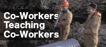 Two field workers standing near a pipe, 'Co-Workers Teaching Co-Workers'